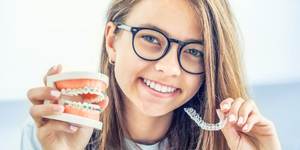 girl with glasses hold teeth model with traditional braces while holding invisaligned in her left